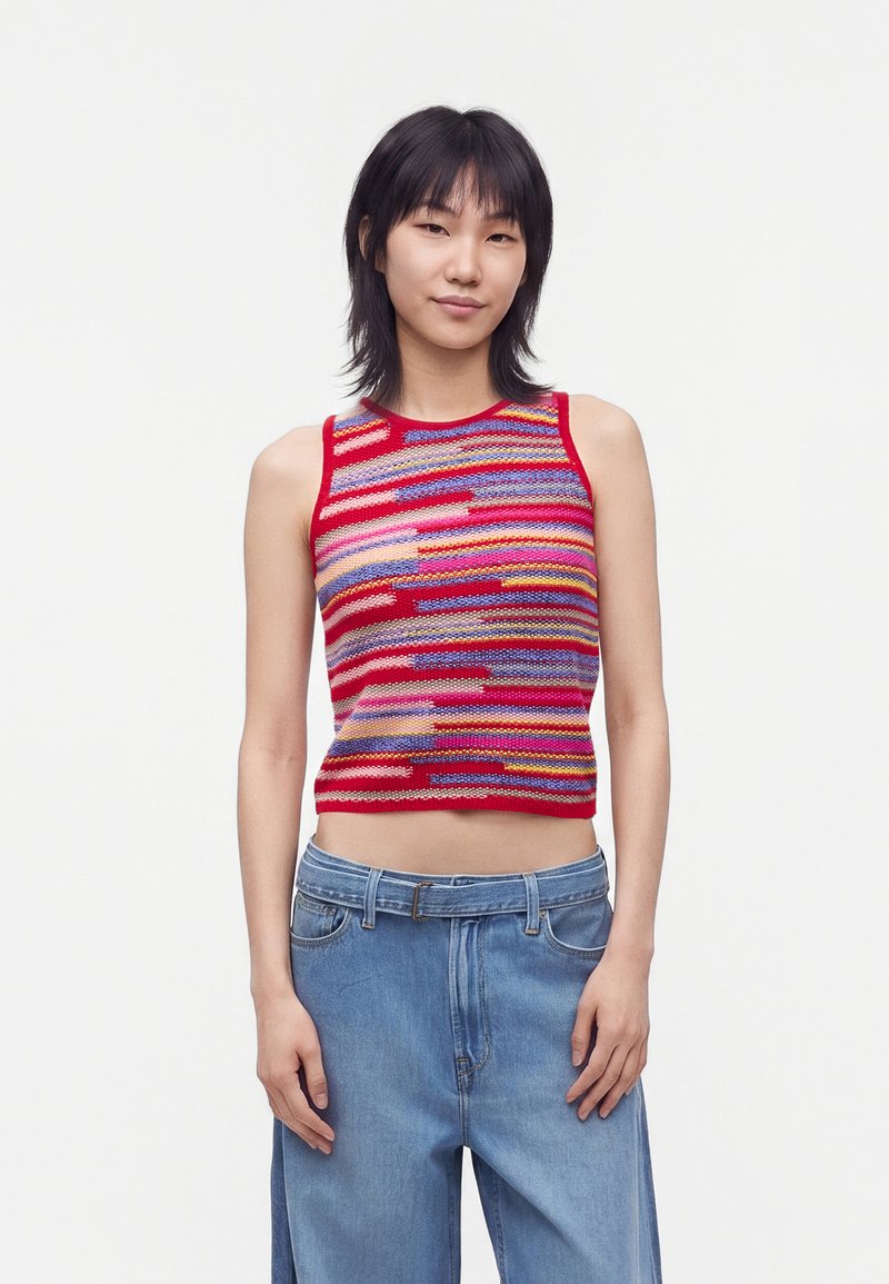 Jeune femme aux cheveux noirs portant un haut en tricot sans manches à rayures colorées et un jean bleu large, debout sur un fond blanc.