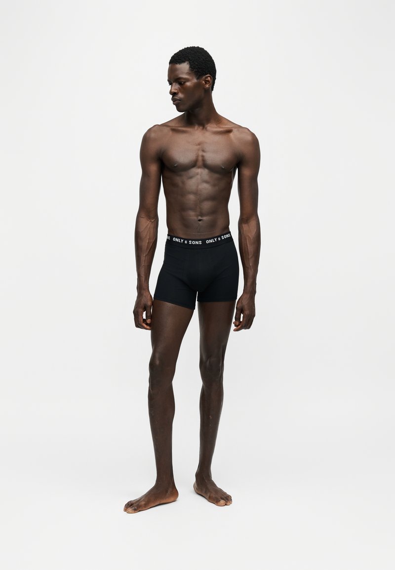 Athletic man standing barefoot wearing black boxer briefs with branded waistband, looking to his left against a plain light background.