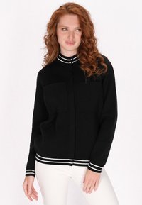 Zwarte cardigan met twee borstzakken, kraag en manchetten in zwart-witte strepen, geribde zoom, zachte stof en een losse pasvorm.
