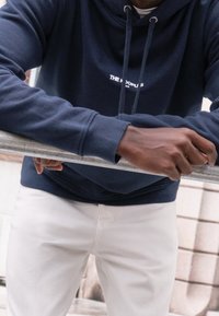 Námořnická modrá mikina s kapucí a přední kapsou, se bílým grafickým textem. Spárovaná s bílými bavlněnými kalhotami. Ruce opřené o kovový zábradlí.