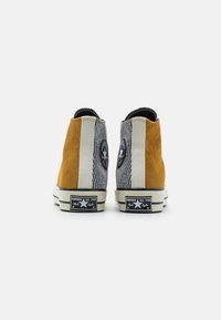 Pár vysokých tenisek Converse z pohledu zezadu, s panelami v hořčicově žluté barvě a černo-bílými pruhy a logovými nášivkami na stranách.