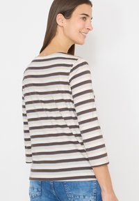 Femme portant un chemisier à manches longues rayé marron, beige et bleu clair, ainsi qu'un jean en denim bleu, souriante et regardant sur le côté.