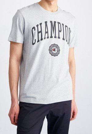 Mężczyzna w jasnoszarej koszulce z dużymi czarnymi literami "CHAMPION" i okrągłym logo marki poniżej, w połączeniu z ciemnymi spodniami.