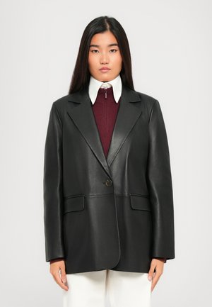 Woman with straight dark hair wears black oversized leather blazer over maroon zip sweater and white collared shirt, paired with white pants.