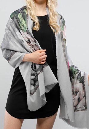 Lyshåret kvinde iført sort kjole med let gråt blomsteret sjal med grønne blade og lyserøde blomster samt tekstbort med "#GORGEOUS".