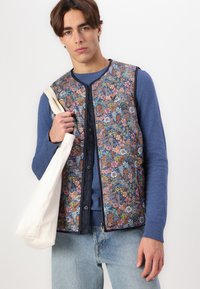 Floral quilted vest with navy trim, colorful patterns of flowers and leaves, worn over a blue sweater, paired with light blue jeans and a tote bag.