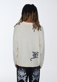 Maglione di lana color crema con una vestibilità rilassata, caratterizzato da un logo ricamato nero sul lato sinistro in basso. Maniche lunghe e polsini a coste.
