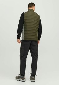 Grüne gesteppte Weste mit hohem Kragen über einem schwarzen Langarmshirt, kombiniert mit schwarzen Cargo-Hosen und grauen Sneakern.