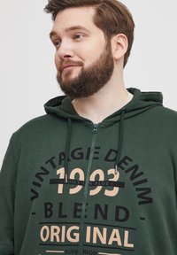 Tamnozelena zip-up dukserica s kenguruvim dzepom, s tiskanim grafičkim motivom u krem i crnoj boji, uključuje vezice i metalne detalje.