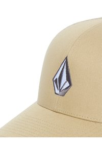 Tan baseballcap met een ronde kroon, gebogen klep en een opvallend geborduurd logo in grijs en zwart aan de voorkant.