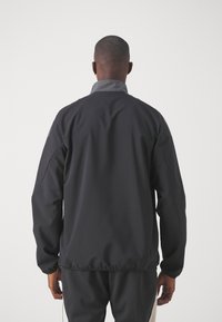 Chaqueta cortaviento negra con cuello gris, que presenta una textura suave y mate y un dobladillo elástico. Diseñada para ofrecer comodidad y movilidad.