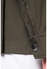 Rękaw oliwkowej kurtki z teksturowanej tkaniny, z zaokrąglonym srebrnym guzikiem i kontrastowym przeszyciem w pobliżu mankietu.