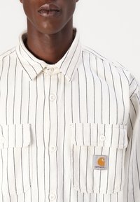 Valge kaelusega särk kitsaste mustade triipudega, kahe esipocket'iga, nööbid ees ja Carhartt'i logo paigatud vasakus taskus.