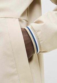 Tejido exterior de color beige con un puño acanalado que presenta rayas en azul, blanco y beige; se muestran en primer plano detalles de la costura del brazo y la manga.