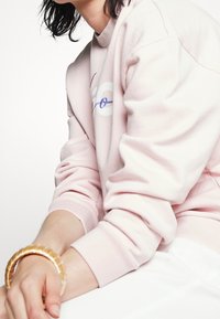 Světle růžová mikina s kulatým výstřihem, na které je vyšívaný text v modré a žluté barvě. Na zápěstí je nošena béžová náramková bangle.