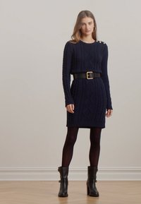 Abito in maglia blu navy con motivi a treccia, maniche lunghe e bottoni sulle spalle. Indossato con una cintura nera e stivali nere con tacco al ginocchio.
