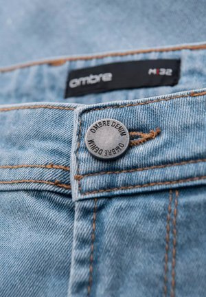Jeans in denim azzurro chiaro con un bottone in argento contrassegnato "OMBRE DENIM," evidenziati da cuciture arancioni, vista della cintura e dell'etichetta visibili.