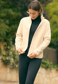 Chaqueta de forro polar color blanco roto con cuello alto y cremallera frontal, llevada sobre una camiseta negra, con leggings negros ajustados. Escenario al aire libre con vegetación.