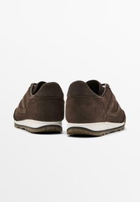 Brown suede athletic shoes with a low profile, rounded heel, white midsole, and textured rubber outsole, featuring stitching details on the upper.