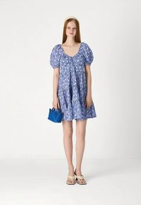 Jovem mulher vestindo um vestido azul com mangas bufantes florais, sandálias bege, faixa de cabeça creme, colar de prata, segurando uma pequena bolsa azul, em pé sobre um fundo branco.