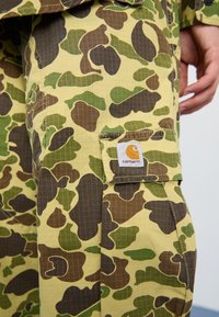 Camo cargo pants in shades of green, brown, and yellow. Features a checked pattern and a patch pocket with a branded label on the side.