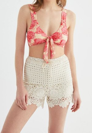 Frau trägt ein rosa Blumen-Crop-Top mit Knoten vorn und hoch taillierte, weiße gehäkelte Shorts vor einem schlichten Hintergrund.