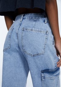 Jeans de mezclilla azul claro con cintura alta, dos bolsillos traseros y un bolsillo lateral de parche. La tela tiene una textura suave y un ligero desvanecimiento.