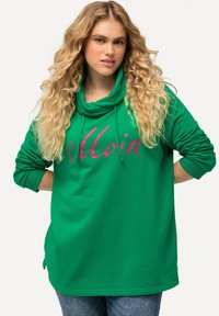 Sudadera verde con cuello vuelto y mangas largas, que presenta un gran texto impreso en rosa que dice "Moin". Tejido suave con un corte relajado.