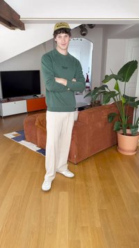 Green sweatshirt, cream wide-leg trousers, and white sneakers. Standing in a room with a brown corduroy couch and tropical plant.
