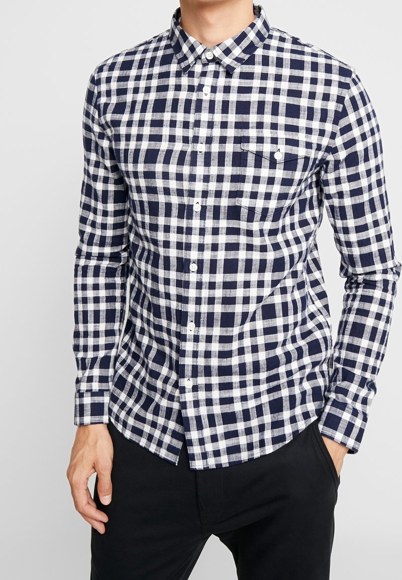 Camicia a quadri blu navy e bianca, con colletto, maniche lunghe e taschino sul petto. Realizzata in tessuto testurizzato, presenta una vestibilità su misura.