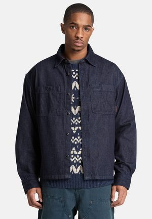 Junger Mann trägt eine dunkle Jeansjacke über einem gemusterten marineblauen und weißen Pullover und steht mit neutralem Gesichtsausdruck vor einem schlichten Hintergrund.