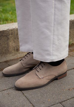 Zapatos de gamuza marrón con cordones y textura lisa, con un diseño cosido y tacón de madera. Combinados con pantalones de corte recto y color claro.