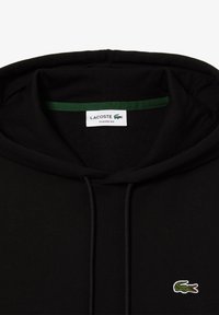 Sweat à capuche noir avec étiquette Lacoste à l’intérieur du col et petit logo crocodile vert brodé sur la poitrine.
