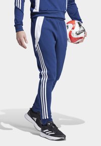 Jūrų kariuomenės sportiniai kelnių su baltais šonais, elastinga juosmens juosta ir siaura prigludimo forma. Laikomi juodi sportiniai bateliai ir raudonai-baltas futbolas.