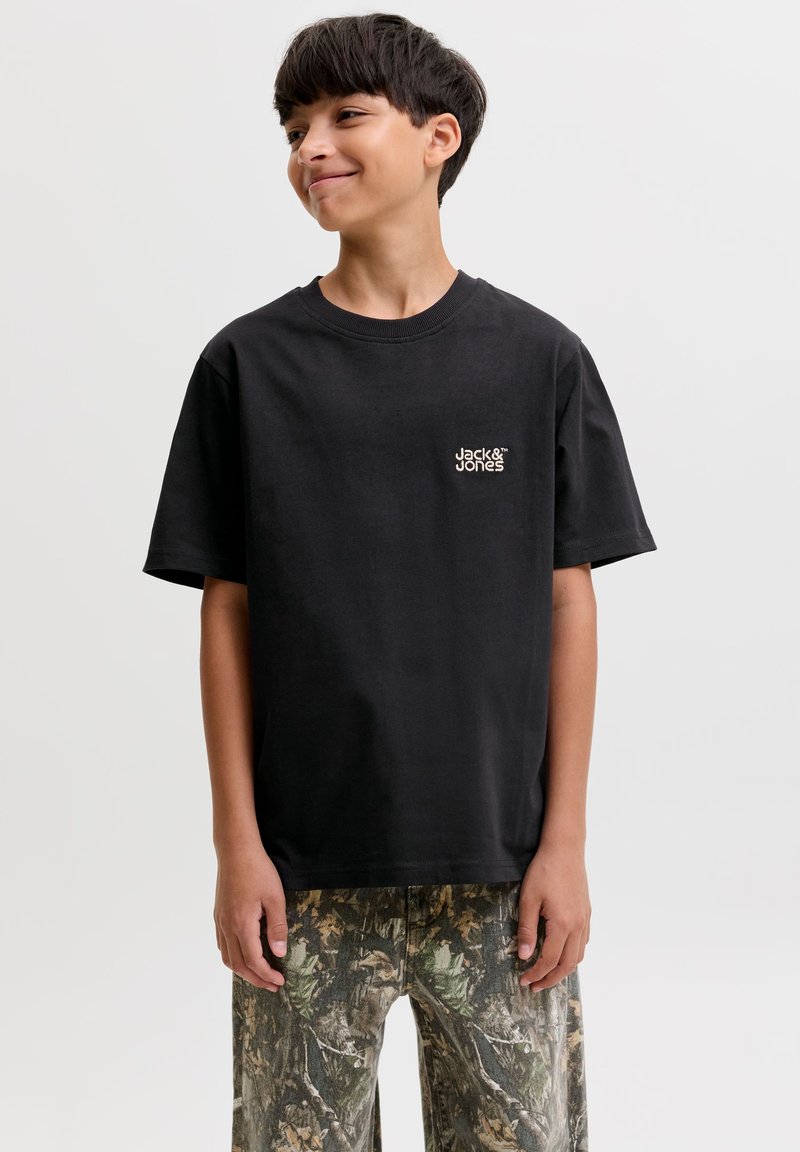 Αγόρι που φοράει μαύρη μπλούζα Jack & Jones και παραλλαγμένο παντελόνι, στέκεται με τα χέρια στα πλάγια, χαμογελώντας και κοιτάζοντας αριστερά.