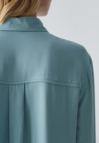 Lichtblauwe, zijden blouse met een shirtkraag, plooien aan de achterkant en een schone, minimalistische uitstraling zonder versieringen.