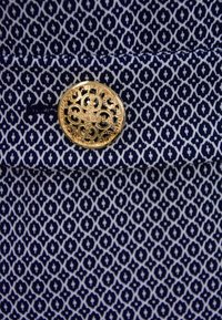 Marinblått tyg med ett vitt geometriskt mönster, prytt med en guldfärgad dekorativ knapp med blommönster. Strukturerad, kompakt väv.