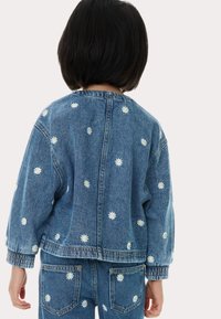 Giacca di jeans con margherite bianche ricamate, caratterizzata da una vestibilità morbida, polsini elasticizzati e cuciture a contrasto, vista da dietro.
