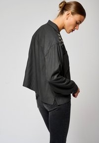 Chemise noire surdimensionnée avec un col boutonné, présentant des accents métalliques décoratifs sur les épaules et un tissu texturé.