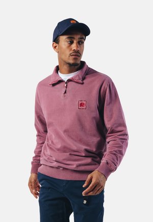 Fleece sweatshirt in stoffige rozenkleur met een ritskraag, voorzien van een vierkante patchlogo op de borst. Gecombineerd met marineblauwe broek.