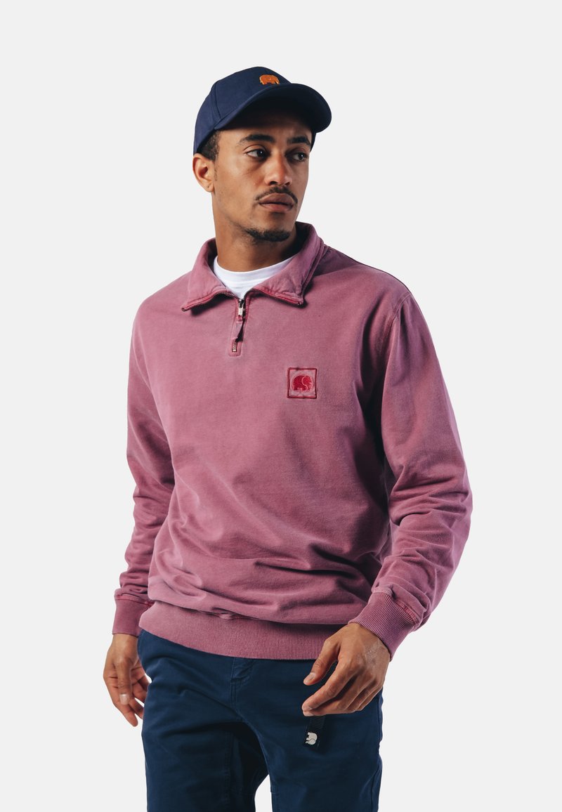 Pull en polaire couleur rose poussiéreux avec un col zippé, arborant un logo en patch carré sur la poitrine. Associé à un pantalon bleu marine.