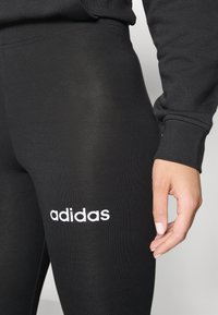 Svarta leggings gjorda av elastiskt material med en slät yta, med en vit "adidas"-logotyp på det nedre benet.
