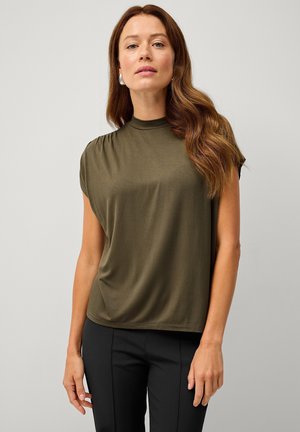 Donkergroene kortebloemige top met een hoge hals, zachte textuur en lichte plooien bij de schouders. Gecombineerd met zwarte broek.