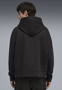 Felpa nera oversize con un ampio cappuccio, spalle scese e polsini a coste. Il materiale sembra morbido e dalla texture piacevole, con un design minimalista.