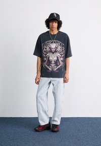 Grafisk svart oversized t-skjorte med detaljert rosa trykk, lyseblå poserende jeans og svarte og røde treningssko, satt mot en nøytral bakgrunn.