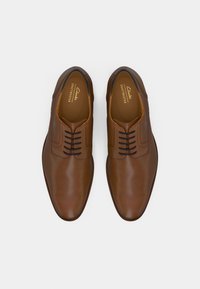 Chaussures habillées en cuir brun avec une texture lisse, bout arrondi et système de laçage. Présente une marque discrète sur la semelle intérieure et un design épuré.