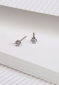 Silberne Steckerohrringe mit einem klaren Edelstein, ausgestattet mit einer Vierkrallenfassung und einem schlanken Steg. Vor einem strukturierten weißen Hintergrund präsentiert.