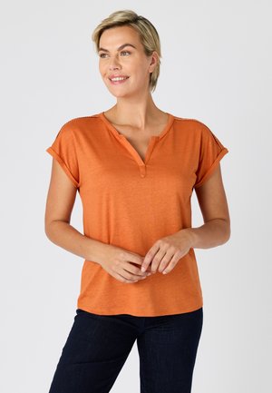 Femme souriante aux cheveux blonds courts portant un haut orange à manches courtes et un pantalon foncé, debout devant un fond clair uni.