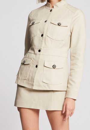 Donna che indossa una giacca beige con bottoni, quattro tasche con pattina e bottoni decorativi, abbinata a una minigonna beige coordinata.