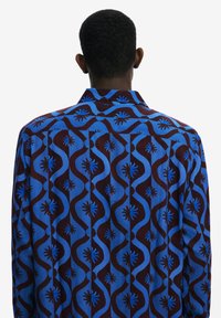 Back view of a shirt featuring a blue and brown abstract pattern, with wavy lines and star-like accents on a textured fabric.
